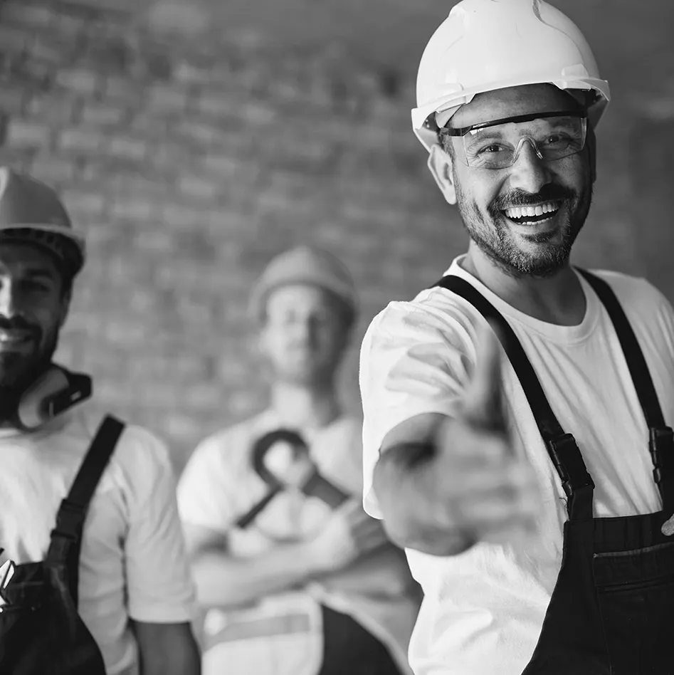 Lachender Bauarbeiter mit Helm und Schutzbrille, im Hintergrund das Team
